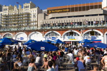 England, East Sussex, Brighton, People sitting under sun shade umbrellas at tables on the promenade outside the Gemini Beach Bar with the De Vere Grand Hotel and The Brighton Centre beyond.