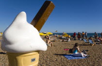 England, East Sussex, Brighton, People sunbathing on the pebble shingle beach with a giant fibreglass model of an ice cream cone in the foreground.