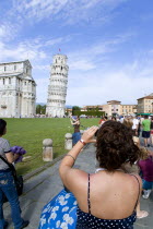 ITALY, Tuscany, Pisa, The Campo dei Miracoli or Field of Miracles.Tourists taking photopraphs of each other pretending to hold up the Leaning Tower or Torre Pendente belltower beyond under a blue sky.