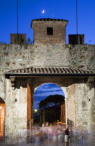 ITALY, Tuscany, Pisa, The Campo dei Miracoli or Field of Miracles.The west entrance gate at dusk with tourists passing through with a half crescent moon in the clear blue sky.