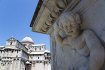ITALY, Tuscany, Pisa, The Campo dei Miracoli or Field of Miracles. The Duomo Cathedral under a blue sky with a marble cherub carved into a plinth in the foreground.