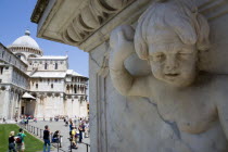 ITALY, Tuscany, Pisa, The Campo dei Miracoli or Field of Miracles with the Duomo Cathedral and tourists under a blue sky with a marble cherub carved into a marble plinth in the foreground.
