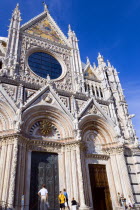 ITALY, Tuscany, Siena, The pink  black and white marble facade of the Duomo cathedral church of Santa Maria Assunta under a blue sky with tourists at the base by the doors.