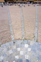 ITALY, Tuscany, Siena, The Piazza del Campo and surrounding buildings with tourists walking in the square. Three lines in flat stone laid into the herringbone brick pattern of the square radiate from...