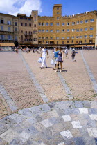 ITALY, Tuscany, Siena, The Piazza del Campo and surrounding buildings with tourists walking in the square. Three lines in flat stone laid into the herringbone brick pattern of the square radiate from...