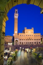 ITALY, Tuscany, Siena, The Torre del Mangia campanile belltower of the Palazzo Publico illuminated at night seen through the archway of the narrow sidestreet leading into the Piazza del Campo with peo...