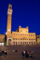 ITALY, Tuscany, Siena, The Torre del Mangia campanile belltower of the Palazzo Publico illuminated at night with people sitting on the ground of the Piazza del Campo.