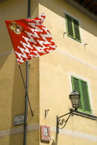 ITALY, Tuscany, San Quirico D'Orcia, Red and white flag and crest of the Castello Quartieri or quarter of the medieval town on the corner of a tellow building in the main square or piazza.