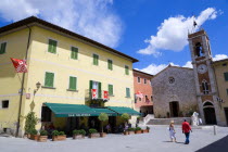 ITALY, Tuscany, San Quirico D'Orcia, Red and white flags of the Castello Quartieri or quarter decorate the Bar Centrale in the main square with a couple walking towards the Church of St Francis or Chi...