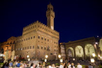 ITALY, Tuscany, Florence, People eating alfresco at a restaurant in The Piazza della Signoria at night with the Palazzo Vecchio and campanile bell tower illuminated beside the Loggia dei Lanzi.