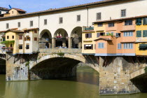 ITALY, Tuscany, Florence, Ponte Vecchio medieval bridge across River Arno with sightseeing tourists beside merchant's shops that line bridge and hang over the water below.