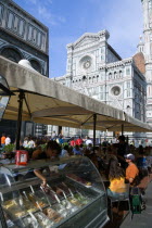 ITALY, Tuscany, Florence, The Neo-Gothic marble west facade of the Cathedral of Santa Maria del Fiore the Duomo with sightseeing tourists people sitting under umbrellas in the shade at a restaurant and a waitress serving ice cream from a counter.