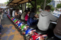 ENGLAND, London, Camden Town, People sitting on small colorful vespa scooters on a line formation under stalls, enjoying the view of Camden Towns canals, while eating their food and other people are w...