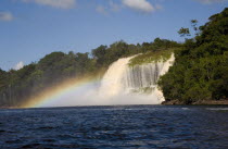 VENEZUELA, Bolivar State, Canaima National Park, Canaima Village, waterfalls feeding into Canaima lake while the humidity of its waters are producing a rainbow effect with blue sky and white clouds.