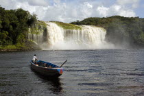 VENEZUELA, Bolivar State, Canaima National Park, Canaima Village, A Pemon tribe man while sailing with his boat at Canaima lake just in front of a big waterfall on a bright day with blue sky and white...