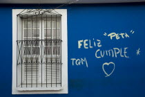VENEZUELA, Bolivar State, Ciudad Bolivar, blue wall with graffiti words in Spanish and the symbol of a heart just next of a closed symmetrical window within a white painted frame, secured with iron ba...