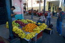 VENEZUELA, Bolivar State, Ciudad Bolivar, Fruit stall with bananas, oranges, apples and other fruits at Ciudad Bolivars main street just by the Orinoco River with people looking at them and talking t...