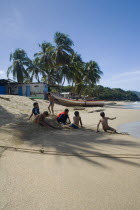 VENEZUELA, Margarita Island, Playa la Galera,  Kids playing on the beach, under the shade of a palm tree just in front of the seawater while other trees, a house and a boat are noticeable at the backg...