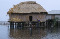 BENIN, Ganvie, Thatched house on stilts in West African lake town,  woman and children on a boat beside it. 