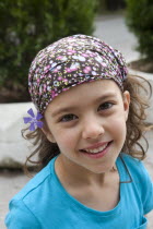 Greece, Macedonia, Thessaloniki, Head and shoulders portrait of a young smiling girl with a purple flower on her ear, wearing a bandanna.
