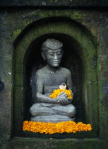 Indonesia, Bali, Ubud, Stone statue of male figure in niche of wall sitting in lotus position holding and surrounded by yellow frangipani flowers.