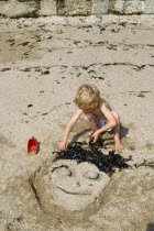 England, East Sussex, Brighton, Young boy making a sand sculpture of a face in the sand on the beach using seaweed and stones.