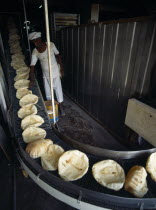 Qatar, Doha City, Baker in bakery standing beside conveyor belt carrying round Arab breads coming out of the oven in the Middle East Gulf State.