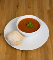 Food, Cooked, Soup, Bowl of tomato and basil soup on a plate with a rustic bread roll on a wooden table surface.