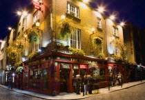 Ireland, County Dublin, Dublin City, Temple Bar Pub illuminated at night with people walking past on the cobbled streets of the area on the south bank of the Liffey River.