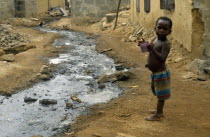 Ghana, Dixcove, Child holding drinking cup standing beside open sewer through street centre.