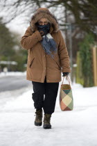 England, West Sussex, Chichester, Middled aged woman in warm clothing carrying shopping bag and walking along icy pavement in winter snow.