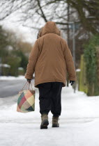 England, West Sussex, Chichester, Middles aged woman in warm clothing carrying shopping bag and walking carefully along icy pavement in winter snow.