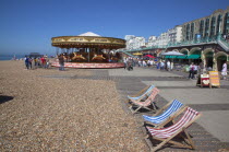 England, East Sussex, Brighton, Deck chairs and carousel on the pebble beach promenade.