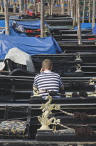 Italy, Veneto, Venice, Gondolier in striped shirt using mobile phone while sitting in one of a line of moored gondolas with back to camera.