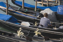 Italy, Veneto, Venice, Gondolier wearing striped shirt using mobile phone sitting in one of a line of moored gondolas with back to camera.