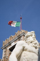 Italy, Veneto, Venice, Centro Storico, Arsenale. Part view of crenellated tower flying Italian tricolour flag with statue of Neptune in foreground against clear blue sky of late summer.