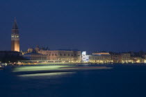 Italy, Veneto, Venice, Giudecca island, Early evening view towards illuminated Campanile of St. Mark with vaporetto water bus to Murano and Burano in blurred motion in foreground with light trails ref...