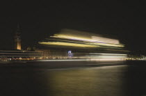 Italy, Veneto, Venice, Giudecca island, Evening view towards illuminated Campanile of St. Mark with vaporetto water bus to Murano and Burano in blurred motion in foreground with light trails reflected...