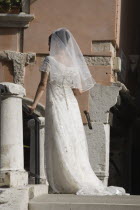 Italy, Veneto, Venice, Bride in white wedding dress and veil, standing on canal bridge in late summer sun, back to camera.