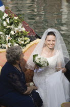 Italy, Veneto, Venice, Smiling bride in wedding dress and veil, carrying bouquet of white roses seated beside mother  on gondola prepared for wedding trip on canal in late summer sunshine.