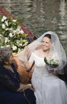 Italy, Veneto, Venice, Smiling bride in wedding dress and veil, carrying bouquet, seated beside mother on gondola prepared for wedding trip on canal in late summer sunshine.