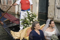 Italy, Veneto, Venice, Cropped shot of gondolier steering decorated gondola with smiling bride in wedding dress and veil, carrying bouquet, and her mother on wedding trip on canal in late summer sunsh...