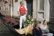 Italy, Veneto, Venice, Gondolier steering decorated gondola carring bride in wedding dress and veil, holding bouquet, and her mother on wedding trip on canal in late summer sunshine.