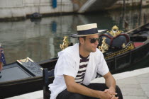 Italy, Veneto, Venice, Centro Storico, Young gondolier in traditional uniform of striped shirt and straw hat with black sash, wearing sunglasses, seated beside moored gondola on canal in late summer s...