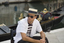 Italy, Veneto, Venice, Centro Storico, Young gondolier in traditional uniform of striped shirt and straw hat with black sash, wearing sunglasses, seated beside moored gondola on canal in late summer s...