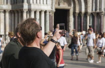 Italy, Veneto, Venice, Centro Storico, St. Marks Square, Tourist takes photograph on mobile phone with tourist crowds and facade of Basilica di San Marco part seen behind.
