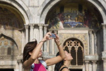 Italy, Veneto, Venice, Centro Storico, St. Marks Square, Female tourist taking photograph on her mobile phone, facade of Basilica di San Marco behind.