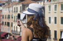 Italy, Veneto, Venice, Young female tourist in white straw hat taking photograph on mobile phone from Rialto bridge looking along Grand Canal.