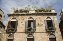 Italy, Veneto, Venice, Building facade with multiple windows, each with small balcony, window box and wooden shutters. Plaque depicting St. George and the Dragon and pots of plants on rooftop above.