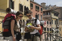 Italy, Veneto, Venice, Two young backpacker tourists wearing sunglasses and carrying rucksacks, rolled up sleeping mats and moneybelts, consulting guide book while standing on canal bridge in Centro S...
